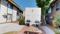 This shared outdoor courtyard serves as a central communal amenity within a multi-unit residential complex. The space is thoughtfully organized into distinct zones designed to encourage outdoor activity and social interaction. A well-maintained lawn area occupies the heart of the courtyard, bordered by established landscaping that includes ornamental shrubs, grasses, and a decorative potted tree, adding visual interest and a natural aesthetic to the surroundings.

Brick herringbone-patterned pathways weave through the space, connecting the various activity areas and providing durable, attractive walkways underfoot. Stone and tile-topped benches are positioned throughout, offering comfortable spots for residents to relax and enjoy the outdoors. A dedicated BBQ area features two freestanding gas grills set against a stucco wall, accompanied by additional seating, making it a practical space for outdoor cooking and entertaining.

The courtyard is enclosed by the surrounding two-story buildings, creating a sheltered, private atmosphere while allowing ample natural sunlight throughout the day. Mature greenery along the building perimeters softens the hardscape and contributes to the overall tranquil ambiance. Concrete pathways near the building facades provide easy access to individual units, and low-profile landscape lighting enhances the space's usability into the evening hours.