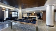 Located at 201 E Gay Street in the heart of West Chester, PA, this single-floor amenity lounge is a thoughtfully designed social space that blends upscale aesthetics with functional entertainment. The centerpiece of the room is a sleek, modern billiards table with a navy felt surface, positioned beneath a striking circular pendant light that draws the eye upward to the rich wood-paneled ceiling above. Structural columns with polished stone-like finishes add an architectural character to the open layout, while recessed lighting provides warm, even illumination throughout. A built-in bookshelf serves as a natural room divider, separating the billiards area from a comfortable lounge zone furnished with plush seating, including an accent chair and a large flat-screen television mounted on a classic brick accent wall. A smaller dining or café-style seating area is visible near the windows, allowing natural light to filter into the space and offering views of the surrounding streetscape. The patterned area rug anchors the space and adds visual texture to the polished floor beneath. Overall, the lounge delivers a refined yet inviting atmosphere, ideal for socializing, relaxing, or casual recreation.