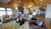 This single-floor space showcases a striking blend of rustic and contemporary design elements throughout its open-concept layout. The living area is anchored by soaring vaulted ceilings finished in warm, natural wood planking, which lend a cabin-inspired character to the space while creating an expansive sense of height and openness. Recessed lighting and stylish pendant fixtures complement the natural warmth of the ceiling, providing layered illumination throughout.

A floor-to-ceiling stacked stone fireplace serves as the focal point of the living area, adding a bold architectural statement and a cozy ambiance. The living space is furnished with a generous sectional sofa, plush accent chairs — including a rich leather armchair — and a substantial dark wood coffee table set atop a patterned area rug, creating a comfortable and well-appointed gathering area.

The open layout flows seamlessly into the dining area and kitchen beyond, where pendant lighting and modern cabinetry are visible. Windows allow natural light to filter in, brightening the neutral-toned walls and enhancing the overall warmth of the interior. The combination of stone, wood, and refined furnishings creates a cohesive, inviting atmosphere well-suited for both relaxed living and entertaining.