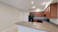 This single-floor space centers around a functional, open-concept kitchen designed for both efficiency and ease of use. The kitchen is anchored by a peninsula counter finished in a neutral gray laminate surface, which houses a double stainless steel sink and provides additional workspace and a casual dining or prep area. Warm medium-toned wood cabinetry lines the walls, offering generous upper and lower storage throughout. Black appliances, including a full-size refrigerator, built-in microwave, and range, create a cohesive and practical cooking environment. The countertops continue along the back wall, maximizing usable workspace. Light beige ceramic tile flooring runs throughout, contributing to a clean, low-maintenance finish. Recessed lighting and a flush-mount ceiling fixture provide layered illumination across the space. White interior doors with simple panel detailing are visible, maintaining a consistent, classic aesthetic. HVAC vents integrated into the ceiling and walls support climate control throughout. The overall ambiance is tidy, practical, and well-organized, suited for everyday living.