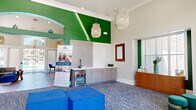 This thoughtfully designed single-floor clubhouse offers a generously proportioned layout that seamlessly integrates a reception area, lounge, dining nook, and full kitchen. The space immediately impresses with its dramatically high vaulted ceilings, bold emerald green accent walls, and crisp white crown molding that together create a vibrant yet polished atmosphere.

The main reception and lounge area serves as the heart of the space, featuring a staffed front desk with warm wood cabinetry, woven pendant light fixtures, and patterned blue-grey carpeting that anchors the open floor plan. A dedicated seating lounge showcases a geometric tile-clad fireplace wall, plush teal velvet armchairs, and coordinated blue ottomans, lending an upscale residential feel. Adjacent to this is an intimate dining nook with a round wood table and mid-century modern chairs, bathed in natural light from large windows overlooking the surrounding greenery.

The kitchen is outfitted with sleek, light wood cabinetry, quartz countertops, a bold hexagonal tile backsplash, and hardwood-style flooring. A kitchenette bar area with a beverage refrigerator provides additional convenience. Throughout the space, curated artwork, decorative mirrors, and natural accents contribute to a cohesive, contemporary design that is both functional and welcoming.