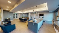 This sophisticated single-floor communal space is thoughtfully designed with a contemporary aesthetic, blending functionality and style across several distinct yet open zones. At the heart of the space sits a striking kitchen island finished in a muted teal-gray tone with a dark countertop, an integrated sink, and elegant cylindrical pendant lights suspended above. Extended counter space runs along a bank of large windows, flooding the area with natural light and offering views to an outdoor terrace beyond.

The dining area features a dark wood table surrounded by deep navy leather chairs beneath a sculptural sputnik chandelier, complemented by a coffered ceiling with warm cove lighting that adds depth and a refined ambiance. Framed artwork adorns the slate-blue accent walls, grounding the space with a curated, gallery-like feel.

A lounge seating area includes plush navy sofas and accent chairs arranged around small side tables, while a trio of gold-framed oval mirrors create a striking decorative focal point along one wall. A separate, more intimate sitting room features leather club chairs, a textured dimensional accent wall, and a modern multi-arm chandelier, offering a quieter retreat. A wall-mounted flat-screen television and additional lounge seating round out the amenities of this well-appointed gathering space.