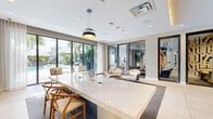 Located at 150 S Bryan Rd in Dania Beach, Florida, this elegantly appointed amenity lounge offers a refined and contemporary atmosphere designed for both productivity and relaxation. The single-floor space is thoughtfully laid out, combining a central living and meeting area with an additional flexible room, all unified by a cohesive modern aesthetic.

The main living area is anchored by a striking white marble conference-style table paired with warm wood-toned wishbone chairs, set atop a plush area rug over large-format tile flooring. A dramatic floor-to-ceiling black marble accent wall serves as a bold focal point, framing a wall-mounted flat-screen television and flanked by warm wood cabinetry. Pendant lighting above the table adds a stylish, intimate touch.

Floor-to-ceiling glass sliding doors and large windows flood the space with natural light and offer sweeping views of a lush outdoor pool terrace lined with tropical palms — bringing the South Florida landscape directly into the interior experience. Sheer drapery softens the light while maintaining the open, airy feel.

A secondary seating area features a pair of curved, upholstered lounge chairs positioned near glass-partitioned private rooms — ideal for informal meetings or focused work. Curated artwork adorns the walls throughout, adding vibrancy and personality to the space's neutral, sophisticated palette.