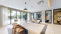Located at 150 S Bryan Rd in Dania Beach, Florida, this elegantly appointed amenity lounge offers a refined and contemporary atmosphere designed for both productivity and relaxation. The single-floor space is thoughtfully laid out, combining a central living and meeting area with an additional flexible room, all unified by a cohesive modern aesthetic.

The main living area is anchored by a striking white marble conference-style table paired with warm wood-toned wishbone chairs, set atop a plush area rug over large-format tile flooring. A dramatic floor-to-ceiling black marble accent wall serves as a bold focal point, framing a wall-mounted flat-screen television and flanked by warm wood cabinetry. Pendant lighting above the table adds a stylish, intimate touch.

Floor-to-ceiling glass sliding doors and large windows flood the space with natural light and offer sweeping views of a lush outdoor pool terrace lined with tropical palms — bringing the South Florida landscape directly into the interior experience. Sheer drapery softens the light while maintaining the open, airy feel.

A secondary seating area features a pair of curved, upholstered lounge chairs positioned near glass-partitioned private rooms — ideal for informal meetings or focused work. Curated artwork adorns the walls throughout, adding vibrancy and personality to the space's neutral, sophisticated palette.
