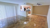 This single-floor residence offers an open and airy layout that connects the living area and kitchen in a cohesive, functional design. The living area features gleaming wood-patterned tile flooring that adds warmth and visual interest throughout the space. Large vertical blinds along one wall allow for ample natural light while providing privacy, and a ceiling fan overhead ensures comfortable airflow year-round.

The kitchen is accessible via a open pass-through layout anchored by a peninsula counter with a dark countertop, creating a natural divide between the cooking and living areas. It is equipped with wooden cabinetry in a warm honey tone, a stainless steel electric range, an over-the-range microwave, and a full-size refrigerator. The kitchen also features a sink integrated into the peninsula, making it a practical and social space for both cooking and entertaining.

The overall ambiance is bright and functional, with neutral wall tones and clean lines that lend a versatile, move-in-ready feel. The open sightlines between the kitchen and living area create a sense of spaciousness, while the consistent flooring throughout reinforces the cohesive design of this well-appointed single-level home.