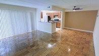 This single-floor residence offers an open and airy layout that connects the living area and kitchen in a cohesive, functional design. The living area features gleaming wood-patterned tile flooring that adds warmth and visual interest throughout the space. Large vertical blinds along one wall allow for ample natural light while providing privacy, and a ceiling fan overhead ensures comfortable airflow year-round.

The kitchen is accessible via a open pass-through layout anchored by a peninsula counter with a dark countertop, creating a natural divide between the cooking and living areas. It is equipped with wooden cabinetry in a warm honey tone, a stainless steel electric range, an over-the-range microwave, and a full-size refrigerator. The kitchen also features a sink integrated into the peninsula, making it a practical and social space for both cooking and entertaining.

The overall ambiance is bright and functional, with neutral wall tones and clean lines that lend a versatile, move-in-ready feel. The open sightlines between the kitchen and living area create a sense of spaciousness, while the consistent flooring throughout reinforces the cohesive design of this well-appointed single-level home.