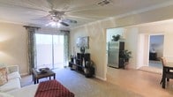 This single-floor residence offers a comfortable and functional layout highlighted by an open-concept design that connects the living area to the kitchen. The living room features plush carpeting, crown molding, and a ceiling fan with an integrated light fixture, contributing to a relaxed and well-ventilated atmosphere. A large sliding glass door along one wall floods the space with abundant natural light while providing access to an outdoor area, framed by patterned drapes that add a decorative touch. The textured ceiling and neutral warm-toned walls create a clean, inviting backdrop throughout. A shelving unit serves as a natural focal point in the living area, offering both storage and display space. The adjacent kitchen is visible through a wide open passageway and features stainless steel refrigerator and light-toned flooring, contrasting pleasantly with the carpeted living space. The transition between rooms is seamless, enhancing the overall sense of openness. The layout feels practical and well-organized, suited for everyday living with a balance of comfort and functionality.