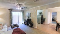 This single-floor residence offers a comfortable and functional layout highlighted by an open-concept design that connects the living area to the kitchen. The living room features plush carpeting, crown molding, and a ceiling fan with an integrated light fixture, contributing to a relaxed and well-ventilated atmosphere. A large sliding glass door along one wall floods the space with abundant natural light while providing access to an outdoor area, framed by patterned drapes that add a decorative touch. The textured ceiling and neutral warm-toned walls create a clean, inviting backdrop throughout. A shelving unit serves as a natural focal point in the living area, offering both storage and display space. The adjacent kitchen is visible through a wide open passageway and features stainless steel refrigerator and light-toned flooring, contrasting pleasantly with the carpeted living space. The transition between rooms is seamless, enhancing the overall sense of openness. The layout feels practical and well-organized, suited for everyday living with a balance of comfort and functionality.