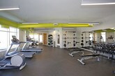 This spacious fitness center is thoughtfully designed to accommodate a full range of workout routines. The interior features durable rubber flooring throughout, white walls, and high ceilings fitted with sleek linear pendant lighting that ensures bright, even illumination across the entire space. A striking design element is the bold yellow exposed ductwork running along the ceiling, adding an industrial-modern aesthetic contrast to the otherwise clean, neutral palette.

The strength training area is well-appointed with a comprehensive dumbbell rack spanning a wide range of weights, multiple weight benches, barbell racks, and cable machine systems. Full-length mirrors line one wall, enhancing the sense of space while allowing users to monitor their form during exercises. Framed fitness-themed artwork decorates the walls, contributing to a motivating atmosphere.

The cardio zone is equally well-equipped, housing elliptical trainers, a rowing machine, and stationary cycling equipment. Large windows and glass doors flood this section with natural light and provide views of the surrounding outdoor environment, creating an open, airy feel. Organized storage shelving near the entrance holds additional accessories and equipment, keeping the space functional and orderly.