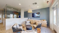 This thoughtfully designed pet amenity space combines functionality with a welcoming, resort-inspired aesthetic. The single-floor layout is finished with polished terrazzo-style flooring that flows throughout, complementing the cool blue-grey and white color palette that defines the space's character.

The lounge area serves as the social heart of the space, furnished with rope-woven chairs, plush cushioned seating, and natural wood accent stools arranged around a central ottoman. A wall-mounted flat-screen TV is positioned above built-in light wood cabinetry, providing both storage and display space. A woven pendant light anchors the seating area, adding warmth to the refined atmosphere.

Adjacent to the lounge, a dedicated grooming station features a sleek, tiled service counter with professional-grade equipment, framed by striking vertical blue tile work that adds a bold design accent. Glass partitions provide visual separation between areas while maintaining an open, airy feel.

The space is adorned with vibrant, oversized dog-portrait artwork displayed prominently along the walls, reinforcing the pet-friendly character throughout. Large glass double doors invite natural light deep into the interior and provide direct access to an outdoor area. A wooden bench near the entry offers a practical spot for pet owners, completing this well-appointed and charming pet amenity lounge.