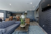 This well-appointed single-floor space combines modern design with warm, inviting elements throughout its open-concept layout. The kitchen is a standout feature, showcasing rich dark espresso cabinetry paired with light stone countertops, a herringbone-patterned tile backsplash, and stainless steel appliances including a French door refrigerator and electric range. A large island with seating provides both workspace and casual dining, while wood-look plank flooring ties the space together seamlessly.

The open floor plan flows from the kitchen into a generous dining area and spacious lounge, where deep charcoal and slate-blue walls create a sophisticated backdrop. Exposed dark brick accents one wall, adding an industrial touch, while a wall-mounted flat-screen television serves as a focal point. The lounge is furnished with a tufted sofa, leather accent chairs, and decorative lamps that contribute to the warm atmosphere.

Multiple sets of large black-framed glass doors line one wall, flooding the interior with natural light and providing direct access to an outdoor patio area, visible beyond and furnished for additional gathering. A ping-pong table and bistro-style café seating add versatile recreational options to the space, making it well-suited for social and communal use.