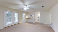 This single-floor residence offers a clean, open layout bathed in natural light. The main living area is generously proportioned, featuring plush carpeting underfoot and a ceiling fan with integrated lighting overhead, ensuring year-round comfort. Neutral tones throughout the space lend a calm, versatile atmosphere well-suited to a variety of furnishing styles.

A pass-through counter connects the living area to the kitchen, creating an open, communicative flow between spaces. The kitchen is equipped with standard appliances, including a microwave and oven range, and features white cabinetry with ample storage. A pendant light fixture adds a subtle decorative touch above the counter.

The living area includes a glass-panel exterior door that invites natural light and provides direct access to an outdoor space, enhancing the sense of openness. A secondary window further brightens the interior.

Visible beyond the main living space is a hallway leading to additional rooms, including a laundry area equipped with a washer and dryer, adding practical convenience to the home. The overall layout is thoughtfully designed to maximize both function and livability, making this a comfortable and well-appointed single-floor residence.
