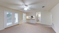 This single-floor residence offers a clean, open layout bathed in natural light. The main living area is generously proportioned, featuring plush carpeting underfoot and a ceiling fan with integrated lighting overhead, ensuring year-round comfort. Neutral tones throughout the space lend a calm, versatile atmosphere well-suited to a variety of furnishing styles.

A pass-through counter connects the living area to the kitchen, creating an open, communicative flow between spaces. The kitchen is equipped with standard appliances, including a microwave and oven range, and features white cabinetry with ample storage. A pendant light fixture adds a subtle decorative touch above the counter.

The living area includes a glass-panel exterior door that invites natural light and provides direct access to an outdoor space, enhancing the sense of openness. A secondary window further brightens the interior.

Visible beyond the main living space is a hallway leading to additional rooms, including a laundry area equipped with a washer and dryer, adding practical convenience to the home. The overall layout is thoughtfully designed to maximize both function and livability, making this a comfortable and well-appointed single-floor residence.