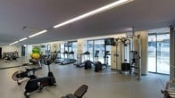 This contemporary fitness center is designed to offer a comprehensive workout experience within a clean, modern environment. The space is laid out across a single floor, featuring a spacious open floor plan with light-toned flooring that creates a bright and energizing atmosphere. Linear LED lighting runs along the ceiling, providing consistent, even illumination throughout the room and complementing the natural light that pours in through a row of large windows along the perimeter wall. These windows also offer views of the surrounding outdoor greenery, enhancing the workout environment. The cardio zone is prominently featured and includes multiple treadmills, elliptical trainers, and stationary bikes, all positioned to take advantage of the natural light and window views. Wall-mounted flat-screen televisions are strategically placed throughout the space, keeping members entertained during their workouts. The strength training area is equipped with a cable machine and multi-functional workout station, suitable for a variety of resistance exercises. Stability balls and additional free weights are also visible, offering flexibility for diverse training routines. The overall ambiance is clean, modern, and professional, making this fitness center a well-appointed facility for a full range of exercise needs.