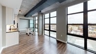 This single-floor unit showcases a compelling blend of industrial and contemporary design throughout its well-organized layout. The most striking feature of the main living area is the wall of large, black-framed floor-to-ceiling windows that flood the space with natural light while offering expansive views of the surrounding cityscape. Exposed concrete columns and ceiling beams complement the rich hardwood flooring below, creating a refined industrial aesthetic. Visible ductwork along the ceiling reinforces the loft-like character of the space.

The kitchen is thoughtfully appointed with white flat-panel cabinetry extending to the ceiling, stainless steel appliances including a range, over-the-range microwave, and refrigerator, complemented by a classic white subway tile backsplash, gray countertops, and matte black hardware for a modern finishing touch.

The full bathroom features a clean, neutral palette with a tub-shower combination, white tiled surround with a built-in niche, a white vanity, and an illuminated LED mirror framed by matte black fixtures and hardware. A separate water closet with neutral gray walls and a matte black towel bar provides additional convenience. Overall, the space delivers a polished, modern urban living experience.