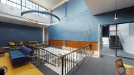Located at 1519 Tower Grove Ave in St. Louis, Missouri, this thoughtfully reimagined historic building masterfully blends original architectural character with modern amenities. The centerpiece of the space is a soaring atrium anchored by a bold circular pendant light that draws the eye upward to clerestory windows flooding the interior with natural light. Distinctive blue-painted walls with deliberately preserved, textured surfaces pay homage to the building's storied past, while warm wood wainscoting and ornate wrought-iron railings add refined detail throughout.

A grand staircase connects the various levels, framed by wood-capped balustrades and open sightlines that enhance the sense of volume and connectivity. Seating areas furnished with navy sofas and accent chairs create inviting lounge spaces ideal for collaboration or relaxation, complemented by dining areas with bold yellow accent furnishings.

Private office spaces feature large multi-pane windows set against exposed brick walls, offering ample natural light and a classic, productive atmosphere with wood wainscoting continuing the building's cohesive aesthetic.

The fitness center is a standout amenity, boasting floor-to-ceiling windows, high ceilings, and a comprehensive array of cardio and strength equipment including treadmills, ellipticals, cable machines, and free weights. An adjoining spin studio with rich wood cabinetry and frosted glass panels provides a dedicated space for cycling workouts.