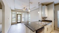 Located at 1460 Distribution Dr in Suwanee, GA, this single-floor residence offers a well-designed open layout that balances functionality and comfort. The kitchen serves as a central hub of the home, showcasing dark granite countertops on a peninsula island, crisp white shaker cabinetry, a tile backsplash, and a full suite of stainless steel appliances including a refrigerator, electric range, microwave, and dishwasher. Track lighting and pendant lights illuminate the space, while tile flooring adds a clean, durable finish to the entry and kitchen areas. The living area is distinguished by a coffered tray ceiling with crown molding, a ceiling fan, and hardwood-style flooring that extends throughout. Multiple windows and a glass-paneled door flood the room with natural light and provide direct access to an outdoor patio area with surrounding greenery. The bedroom features plush carpet, a ceiling fan, and a bright window with outdoor views, creating a calm and comfortable retreat. The bathroom is finished with granite vanity counters, tile flooring, and a full soaking tub. A stacked in-unit washer and dryer add everyday convenience, and a dedicated storage closet with wire shelving provides additional organizational space.