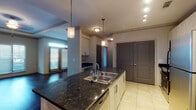 This single-floor residence offers a thoughtfully designed layout with a seamless blend of style and functionality. The open-concept kitchen serves as the heart of the home, showcasing dark granite countertops, a double-basin stainless steel sink set within a central island, and white cabinetry accented by a classic subway tile backsplash. Stainless steel appliances, including a range and refrigerator, complement the space, while tile flooring and pendant lighting add to its polished character. The kitchen flows naturally into the living area, which features rich, dark hardwood floors, an elegant tray ceiling with crown molding, and large windows fitted with blinds that allow for abundant natural light. A secondary bedroom offers plush carpeting, a ceiling fan, and a window with an outdoor view, while another carpeted room provides access to a closet and additional storage. The bathroom is finished with granite countertops, white cabinetry, large-format tile flooring, and a full tub-shower combination with clean white tile surround. Throughout the space, two-panel gray interior doors provide a consistent, modern aesthetic. The entry area features tile flooring and a bold gray front door, creating a welcoming first impression.
