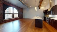 Located at 826 West Sprague Avenue in downtown Spokane, this urban loft seamlessly blends historic character with contemporary finishes. The open-plan living and kitchen area is defined by soaring ceilings, exposed timber beams, and original brick accent walls that speak to the building's storied past. Dramatic arched windows flood the space with natural light while framing views of the surrounding cityscape.

The chef's kitchen is a standout feature, equipped with rich dark cabinetry, white quartz countertops, a subway tile backsplash, stainless steel appliances including a gas range, built-in microwave, and a large center island with an integrated sink — all illuminated by sleek pendant lighting. Warm hardwood flooring runs throughout the main living areas, while exposed ductwork adds to the industrial aesthetic.

The bedroom offers plush carpeting, additional arched window views, and direct access to a well-appointed bathroom featuring large-format tile flooring, a soaking tub with tiled surround, a white quartz vanity, and a heated towel rack. A dedicated utility area houses a Carrier HVAC system. This thoughtfully designed loft delivers an exceptional urban living experience rooted in architectural authenticity.