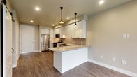 This well-appointed single-floor unit offers a thoughtfully designed layout with modern finishes throughout. The open-concept kitchen serves as the heart of the space, showcasing crisp white shaker cabinetry, a striking granite peninsula that provides additional seating and prep space, and a classic subway tile backsplash. Two stylish black pendant lights hang above the peninsula, complementing the recessed lighting overhead. Stainless steel appliances — including a gas range, built-in microwave, farmhouse sink with dishwasher, and full-size refrigerator — complete the kitchen's functional and contemporary aesthetic. Warm wood-look flooring flows seamlessly throughout the entire unit, tying each space together with a cohesive, inviting feel. The bathroom is finished with large-format tile on the tub surround, a granite-topped vanity with modern cabinetry, and recessed lighting for a clean, polished look. The bedroom features a distinctive sliding barn door, adding a touch of character to the otherwise neutral, modern palette. Neutral wall tones and clean white trim throughout create a bright, airy atmosphere well-suited for comfortable everyday living.