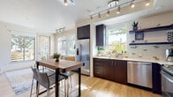 This thoughtfully designed single-floor residence offers a cohesive contemporary aesthetic carried throughout every room. The open kitchen is a standout feature, showcasing rich dark espresso cabinetry, stainless steel appliances — including a French door refrigerator, gas range, microwave, and dishwasher — complemented by a neutral tile backsplash and light quartz countertops. A built-in high-top dining table with bar-height seating sits adjacent to the kitchen, making the space ideal for casual meals and entertaining. Light hardwood flooring flows seamlessly throughout the main living areas, enhancing the bright, airy atmosphere created by an abundance of large windows dressed with horizontal blinds that frame greenery views from multiple angles. The living room offers generous natural light and direct access to an outdoor area, with a wall-mounted television and built-in media console adding functional style. The bedroom is a calm, well-proportioned retreat with neutral tones and soft lighting. Two bathrooms mirror the home's design language, each featuring floating dark-wood vanities, framed mirrors, and light wood-style flooring. The attached garage provides substantial additional utility space, complete with its own window and automatic door opener, rounding out this well-appointed and move-in-ready residence.