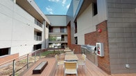 Located at 1212 S East Ave. in Baltimore City's Canton neighborhood, this modern single-floor residence is part of a contemporary multi-story building characterized by a striking mix of exposed brick, metal panel cladding, and clean architectural lines. The building's design centers around a shared outdoor courtyard deck, which serves as a standout communal amenity. The expansive wood-composite deck offers outdoor seating and dining arrangements, a stainless steel built-in grilling station, and open-air views of the surrounding building structure, with glass-panel railings providing an unobstructed sightline to the landscaped courtyard below. The deck is framed by multi-level balconies and walkways, creating a dynamic and social outdoor environment open to natural light and sky. Inside, the bathroom is thoughtfully designed with neutral tones and modern finishes throughout. The vanity features warm maple cabinetry, granite countertops, an undermount sink, and a large mirror, complemented by a natural light-admitting window. The tiled flooring provides a durable, cohesive aesthetic. A separate glass-enclosed shower is finished with large-format stone-look tiles and includes a built-in bench and chrome hardware, offering a spa-like experience. Accessibility features such as grab bars are also present. The unit transitions seamlessly to carpeted living areas through clean white-paneled doorways.