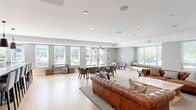 Located at 205 W Touhy Ave in Park Ridge, IL, this single-floor space offers a well-designed open-concept layout that seamlessly combines a fully equipped kitchen and a generous living area. The kitchen is a standout feature, showcasing white shaker-style cabinetry with glass-front uppers, a soft blue tile backsplash, and a large two-tone island with a crisp white countertop and slate-blue base. Multiple pendant lights hang above the island, which provides bar seating for several guests. Stainless steel appliances, including a French door refrigerator and built-in microwave, complete the well-appointed cooking space. The living area is warm and inviting, anchored by a classic fireplace with a white mantel and tile surround. Light hardwood flooring runs throughout the entire space, enhancing its bright, airy feel. Multiple windows dressed with white blinds flood the room with natural light, while a built-in window bench offers a cozy nook. Crown molding and recessed lighting add refined architectural detail to the neutral gray and white walls. A set of glass doors provides direct access to an outdoor patio area, further extending the functionality and openness of this thoughtfully designed communal space.