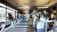 This contemporary fitness center offers a thoughtfully designed, single-floor layout that balances functionality with a sleek, modern aesthetic. The space is anchored by an expansive cardio zone, featuring rows of treadmills, elliptical trainers, and stair climbers equipped with individual digital displays. Industrial-style ceilings with exposed ductwork are complemented by a combination of pendant lighting and linear fixtures, creating a bright and energizing atmosphere throughout. Large windows along one wall invite natural light and provide a connection to the outside, enhancing the open and airy feel of the space. The flooring transitions between dark rubberized surfaces in the cardio zone and wood-look planks in adjacent areas, adding visual warmth and clearly delineating workout zones. Further into the space, a dedicated strength and functional training area is visible, equipped with additional machines and free weights. A feature wall with a graphic or mural adds a dynamic visual focal point, while a mounted display screen supports entertainment or instructional use. The overall ambiance is clean, modern, and motivating, making this fitness center a well-rounded facility suited for a wide range of exercise needs.