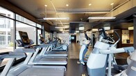 This contemporary fitness center offers a thoughtfully designed, single-floor layout that balances functionality with a sleek, modern aesthetic. The space is anchored by an expansive cardio zone, featuring rows of treadmills, elliptical trainers, and stair climbers equipped with individual digital displays. Industrial-style ceilings with exposed ductwork are complemented by a combination of pendant lighting and linear fixtures, creating a bright and energizing atmosphere throughout. Large windows along one wall invite natural light and provide a connection to the outside, enhancing the open and airy feel of the space. The flooring transitions between dark rubberized surfaces in the cardio zone and wood-look planks in adjacent areas, adding visual warmth and clearly delineating workout zones. Further into the space, a dedicated strength and functional training area is visible, equipped with additional machines and free weights. A feature wall with a graphic or mural adds a dynamic visual focal point, while a mounted display screen supports entertainment or instructional use. The overall ambiance is clean, modern, and motivating, making this fitness center a well-rounded facility suited for a wide range of exercise needs.