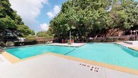 This outdoor pool area offers a well-designed recreational retreat centered around a large, clearly marked swimming pool with a shallow end depth of 3 feet 6 inches. The pool's bright turquoise water is complemented by a wide, light-colored concrete sun deck that surrounds the pool on multiple sides, providing ample space for lounging and movement. A central island feature within the pool area adds visual interest and serves as a gathering point, equipped with orange bar-height stools for casual poolside socializing. Lounge chairs are arranged along the deck, offering comfortable spots to relax and take in the surroundings. A red shade sail provides a shaded seating area on one side of the deck, ideal for those seeking relief from direct sun. The entire area is enclosed by a black metal perimeter fence, ensuring safety and privacy. A backdrop of mature, full-canopy trees frames the space beautifully, offering natural shade and a lush green aesthetic that enhances the outdoor ambiance. The combination of open sky, mature landscaping, and thoughtfully arranged amenities makes this pool area a practical and inviting outdoor space suited for both leisure and light recreation.