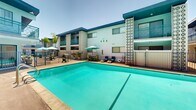 Located at 2635 Milton Ave in Fullerton, California, this two-story mid-century property showcases a distinctive retro architectural style characterized by bold geometric lattice screens, clean horizontal lines, and a crisp white-and-teal exterior color palette. The building's design is immediately striking, with ornamental breeze-block panels flanking the structure and teal-accented railings running along the upper-level balconies, which overlook the central outdoor area.

The heart of the property is a generously sized outdoor courtyard centered around a sparkling swimming pool with vivid turquoise water. The pool deck is well-appointed with lounge chairs, shaded umbrella seating, and comfortable outdoor furniture, creating an inviting space for relaxation. A fenced perimeter provides safety and definition to the pool area, while lush tropical plantings add splashes of greenery throughout the courtyard.

A teal open-riser staircase connects the ground floor to the upper level, reinforcing the building's mid-century modern aesthetic. Ground-floor units open directly onto the pool deck, while upper-floor units enjoy private balcony access with views over the courtyard. The overall ambiance is relaxed and resort-like, blending Southern California sunshine with a thoughtfully preserved retro character that gives this Fullerton property a memorable and distinctive presence.