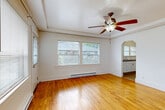 Located at 2630 NW Vaughn St in Portland, Oregon, this single-floor unit offers a well-organized layout comprising a living room, kitchen, bathroom, and hallway. The living room is a standout feature, showcasing warm hardwood floors that extend throughout the space, a ceiling fan with light fixture, and a tray ceiling detail that adds architectural character. Multiple windows along the exterior walls flood the room with natural light, offering views of the surrounding neighborhood's greenery. Baseboard heating runs along the walls, providing efficient climate control. A charming arched doorway connects the living room to the kitchen, adding a classic design touch. The kitchen is equipped with white cabinetry, rich wood-toned countertops, white tile backsplash, a double-basin stainless steel sink, a white electric range, and a full-size refrigerator. Vinyl flooring and multiple windows make the kitchen feel bright and practical. The bathroom features full white tile surround on the tub/shower combination, with a window providing natural ventilation. The overall aesthetic is clean and classic, with white walls and traditional architectural details that reflect the character of Portland's established Northwest neighborhoods.