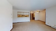 This single-floor space is designed with an open-concept layout that seamlessly connects the kitchen and living area, creating a cohesive and functional environment. The kitchen features a pass-through counter with white upper cabinetry trimmed in warm wood tones, offering both practical storage and a visual connection to the main living space. A refrigerator and cooktop are visible within the kitchen, providing essential appliances in a compact, efficient arrangement.

The living area is generously sized, finished with neutral wall-to-wall carpeting that contributes to a warm and understated ambiance. White walls and a smooth ceiling keep the space feeling bright and clean. A ceiling fan with a light fixture is centrally positioned overhead, providing both air circulation and ambient lighting.

Toward the rear of the space, a wood-toned door leads to what appears to be a private room or bathroom, with interior lighting visible through the doorway. Dark wood baseboards run consistently throughout, offering a subtle contrast to the light-colored walls and flooring. Overall, the layout is practical and straightforward, suited for comfortable everyday living.