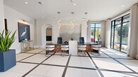This elegant single-floor space serves as a contemporary residential leasing and amenity lobby, thoughtfully designed to make a strong first impression. The entry foyer welcomes visitors with expansive floor-to-ceiling black-framed glass doors and windows that flood the interior with natural light, casting geometric patterns across the striking large-format white tile floors accented with dark inlay lines.

The main lobby area features a prominent leasing desk with a woven wood base and dark stone countertop, complemented by modern white guest chairs and cascading globe pendant lighting overhead. Curated artwork, including a standout egret painting and a backlit textural wall installation, add artistic depth throughout the space. Elegant archways frame transitions between areas, lending a classic architectural touch to the otherwise modern interior.

Adjacent to the lobby, a spacious corridor connects to a mailroom and package area, highlighted by warm wood flooring and a cluster of industrial-style pendant lights above. A decorative metal lattice screen partition adds visual interest and defines spaces without sacrificing openness. A comfortable seating nook near the entrance, with upholstered chairs and views of lush landscaping, offers a welcoming retreat. The overall ambiance is polished and inviting, blending contemporary furnishings with refined architectural details.