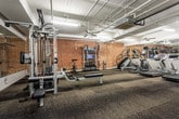 This fitness center in Atlanta, Georgia, offers a comprehensive workout environment characterized by an industrial aesthetic. Exposed concrete ceilings with visible ductwork, conduit, and ceiling fans run throughout the space, contributing to an open, loft-like atmosphere. The flooring consists of durable, multi-tone carpet tiles suited for high-traffic exercise use.

The cardio zone houses multiple Star Trac treadmills and a stair climber, each equipped with individual monitors, as well as an elliptical machine. Large floor-to-ceiling mirrors line the walls of this section, enhancing the sense of space and allowing users to monitor their form.

The strength training area features a multi-station cable machine, a dedicated free weight section with a full rack of dumbbells, adjustable benches, and a stability ball. A functional training station along the exposed brick wall includes a RAX rack system loaded with kettlebells, battle ropes, resistance bands, and a media screen for guided workouts.

The exposed brick walls on one side of the facility provide a distinct urban character, contrasting with the white-painted walls and mirrored surfaces elsewhere. Ceiling fans and fluorescent lighting ensure consistent airflow and visibility throughout the entire fitness space.