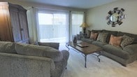 This single-floor residential space offers a comfortable and welcoming layout designed for everyday living. The living room serves as the central gathering area, featuring wall-to-wall carpeting that adds warmth underfoot and a soft, neutral color palette throughout. A large sliding glass door dominates one wall, dressed with vertical blinds and sheer curtains that allow for adjustable natural light while providing a direct connection to an outdoor patio area. This generous source of natural light brightens the entire room, creating an open and airy atmosphere. The space is furnished with plush, cushioned sofas arranged around a sturdy wood-and-wrought-iron coffee table, offering both style and functionality. A classic table lamp provides warm ambient lighting for evening use. The walls are finished in a muted, neutral tone that complements the earthy furnishings and contributes to the overall relaxed ambiance. The room's layout is practical and spacious, allowing for comfortable movement and flexible furniture arrangement. Overall, this single-level space presents a cozy, well-lit living environment with a seamless indoor-outdoor connection that enhances its livability.