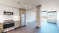 This well-appointed one-bedroom, one-bathroom unit offers a thoughtfully designed open-concept layout that seamlessly connects the kitchen and living areas. A defining architectural feature throughout the space is the exposed concrete columns and ceiling elements, which lend an industrial-modern character that contrasts beautifully with the clean white walls and warm wood-tone finishes.

The kitchen is outfitted with a full suite of stainless steel appliances, including a gas range, microwave, dishwasher, and refrigerator. Cabinetry combines glossy white upper cabinets with wood-grain lower cabinets, complemented by white quartz countertops. Dark hardwood-style flooring runs throughout the main living areas, adding warmth and continuity.

The living area is bathed in natural light through expansive floor-to-ceiling windows that frame impressive views of the waterfront, a river, and the surrounding cityscape — a standout feature of this unit. The bedroom continues this theme with large sliding glass doors opening to equally striking panoramic views, and is carpeted for comfort. It is served by a dedicated walk-in closet with built-in shelving and hanging rods.

The bathroom features a wood-grain vanity with a white quartz countertop, a wide mirror with accent lighting, and clean tile flooring, completing this modern and cohesive living space.