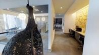 This single-floor space showcases a thoughtfully designed interior that blends artistic expression with functional design. A dramatic sculptural installation — an intricately carved dark pendant piece — serves as an immediate focal point upon entry, setting a bold, artistic tone for the entire space. The corridor leading through the space is finished with neutral carpeting and crisp white walls, lending a clean, gallery-like quality to the layout.

Recessed ceiling lighting runs throughout, complemented by distinctive globe pendant lights visible through a glass-partitioned area, which add warmth and visual interest. The glass partition allows borrowed light to filter through while maintaining a sense of openness and connectivity between zones.

Along one wall, a built-in workstation with dark cabinetry provides a sleek, integrated workspace complete with ergonomic seating and a monitor setup. Adjacent to this, a textured wall treatment adds depth and dimension, contrasting beautifully with the otherwise smooth white surfaces.

Further into the space, a meeting or conference area is visible, furnished with modern chairs and a long table beneath the globe pendants. The overall ambiance is polished and contemporary, with carefully chosen design elements that balance creativity and professionalism.