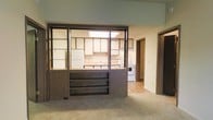 This single-floor residence offers a practical and functional layout designed for comfortable everyday living. The standout architectural feature is a bold, dark-framed open shelving unit that serves as a room divider between the carpeted living area and the kitchen, providing both visual separation and display storage while maintaining an open, connected feel throughout the space.

The kitchen is well-equipped with white appliances, including a refrigerator and a stove, complemented by light wood cabinetry that lines the walls. A skylight overhead floods the kitchen with natural light, brightening the space and adding an airy quality. The countertops and sink are positioned conveniently along the kitchen perimeter, making the layout efficient for daily use.

The living area features wall-to-wall carpeting that lends a warm, comfortable atmosphere, while the kitchen transitions to smooth, light-toned vinyl or laminate flooring for easy maintenance. A hallway visible from the main living space leads to additional rooms, offering clear circulation throughout the home. Neutral wall tones and wood-accented trim throughout contribute to a cohesive, understated aesthetic that provides a versatile backdrop for a variety of interior styles.