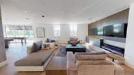This thoughtfully designed amenity space spans multiple levels connected by open staircases with sleek black metal and glass railings, creating a sense of vertical openness throughout. The main lounge area is anchored by a wide linear fireplace set within a dark wood-paneled accent wall, flanked by large flat-screen displays and complemented by plush neutral-toned sofas arranged over a richly patterned area rug atop warm hardwood floors. Natural light pours in through generously sized windows, offering views of lush greenery beyond. A dedicated meeting and dining area features a round wood-top table surrounded by modern blue accent chairs beneath a striking circular pendant light, with a textured dark wood feature wall providing a refined backdrop. Adjacent to this, a reading or lounge nook is defined by elegant slatted wooden screens that filter light beautifully, furnished with rich brown leather wingback chairs. A decorative lattice-patterned partition adds visual interest and subtly separates zones throughout the space. A full indoor golf simulator room, finished in deep matte black, provides an immersive recreational experience. Upper-level mezzanines house a fitness center enclosed in floor-to-ceiling glass walls, offering panoramic views of the surrounding landscape. The overall ambiance is polished, modern, and versatile.