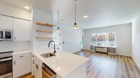 This single-floor unit showcases a clean, modern design defined by a bright, open-concept layout that seamlessly connects the kitchen and living areas. Light wood-tone vinyl plank flooring runs throughout, lending warmth and continuity to the space.

The kitchen is a standout feature, offering white flat-front cabinetry with matte black hardware, quartz countertops, a subway tile backsplash, and stainless steel appliances including a range, microwave, and refrigerator. A peninsula with an undermount sink and matte black pull-down faucet provides additional prep space and casual seating potential. Live-edge wooden floating shelves add a natural, artisan touch, while pendant lighting overhead creates an inviting atmosphere.

The living area is generously proportioned, bathed in natural light through wide windows fitted with blinds. A built-in shelf unit beneath the windows provides practical storage alongside an in-wall HVAC unit.

The bedroom is well-sized and bright, featuring large windows and the same wood-tone flooring. The bathroom maintains the unit's modern aesthetic with a white quartz vanity, undermount sink, matte black fixtures, a large mirror, and a tub-shower combination, all finished in crisp white.