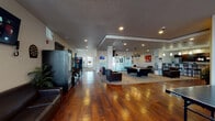 This single-floor amenity space is thoughtfully designed to serve as a multifunctional recreational and social hub. Rich hardwood flooring extends throughout, tying together the various activity zones and lending warmth to the overall aesthetic. Recessed lighting illuminates the open layout, complemented by pendant fixtures that highlight key gathering areas.

The game room is a standout feature, centered around a full-size billiards table beneath a classic hanging lamp, accompanied by a ping-pong table nearby. A dartboard mounted on the wall adds another layer of entertainment options. Vending and snack machines are conveniently positioned throughout the space for easy access.

Multiple seating arrangements featuring dark leather sofas and armchairs are distributed across the lounge areas, creating comfortable zones for relaxation and socializing. Wall-mounted flat-screen televisions are present in several areas, enhancing the communal viewing experience.

The kitchen area is well-equipped with dark cabinetry, granite-style countertops, a full refrigerator, a coffee maker, and ample counter space, making it suitable for group gatherings. Track and recessed lighting brighten this functional area. Glass-paneled partitions provide subtle separation between zones while maintaining the open, connected feel of the entire space.