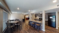 Located on S Gessner Road in Houston, TX, this single-story community space is designed to serve as a functional and comfortable gathering area. The layout follows an open floor plan concept, with distinct zones flowing seamlessly into one another across rich, wood-look plank flooring that runs throughout the entire space.

The kitchen area is well-equipped with dark gray cabinetry, light quartz-style countertops, black appliances including a full-size refrigerator, microwave, and dishwasher, along with a sink and ample counter space. A peninsula with bar-height seating and wooden stools separates the kitchen from the adjacent dining area, which features multiple dining tables and chairs positioned near large windows and a glass door that allow natural light to fill the space and provide views to the exterior.

The lounge area offers comfortable seating arrangements with upholstered sofas and armchairs grouped around wall-mounted flat-screen televisions. A ceiling fan provides additional comfort. A designated children's nook is also incorporated into the layout, featuring colorful furniture and play items. Recessed lighting throughout ensures consistent illumination, while the neutral wall tones and clean finishes contribute to an organized and welcoming overall ambiance.