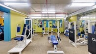Located at 2323 Lake Shore Blvd W in Toronto, this fitness facility offers a well-appointed exercise room designed to support a comprehensive workout experience. The space is divided into distinct functional zones, each catering to different training needs. The cardio area features multiple treadmills and elliptical machines, complemented by a wall-mounted television for entertainment during workouts. The strength training zone is outfitted with a robust selection of cable machines, weight benches, and multi-use resistance stations from brands including Pulse, providing users with ample options for full-body conditioning. Stability balls and additional accessories round out the equipment available. The interior is characterized by vibrant yellow and green accent walls paired with bold teal-blue doors and columns, giving the space a lively, energetic atmosphere. Fluorescent lighting illuminates the facility throughout, while exposed overhead piping adds an industrial character to the ceiling. Large mirrors line sections of the walls, enhancing the sense of openness and allowing users to monitor their form. Durable carpet tile flooring covers the exercise areas, providing comfort underfoot. The facility is accessed through keyed entry doors and includes wall-mounted sanitation dispensers, supporting a clean and functional environment for residents.
