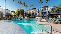 Located at 9680 W Northern Ave in Peoria, Arizona, this outdoor amenity space delivers a resort-inspired experience designed for relaxation and recreation under the Arizona sun. The centerpiece of the space is a generously sized, freeform swimming pool with vivid turquoise water, accented by decorative tile inlays and framed by a wide, open sun deck. Adjacent to the pool sits a circular spa, seamlessly integrated into the overall design and ideal for unwinding year-round.

The pool deck is well-appointed with rows of teal lounge chairs and coordinating sun umbrellas, providing ample shaded and open-air seating options. Decorative bowl-style water features add an elegant architectural touch at the pool's edge. Towering palm trees rise throughout the space, lending a tropical ambiance while offering natural shade and visual interest against the clear blue Arizona sky.

A separate outdoor lounge area offers a colorful circular seating piece with vibrant accent pillows beneath a shade structure, positioned near a gated entry surrounded by neatly trimmed hedges and mature shade trees. The surrounding multi-story building features contemporary architecture with private balconies, large windows, and a modern gray and blue exterior that complements the outdoor setting. The overall atmosphere is vibrant, welcoming, and tailored for outdoor living.