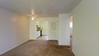 This single-floor space presents a straightforward, functional layout with a neutral color palette throughout. The main living area is finished with wall-to-wall beige carpeting, creating a warm and cohesive feel underfoot. A flush-mount ceiling light fixture provides overhead illumination, and the smooth white walls offer a clean, understated backdrop throughout the space.

Visible from the living area, the kitchen features white cabinetry along the walls, dark countertops, and a black appliance, providing a practical cooking space. The kitchen's flooring transitions from the carpeted living area, and the room benefits from natural light filtering through a nearby window.

A distinctive louvered door is centrally positioned, providing access to an additional area, likely a closet or utility space. To the right, an open passageway leads to a further room, which also receives natural light from a window, suggesting it may serve as a bedroom or secondary living space.

The textured ceiling adds a traditional finish throughout, and the overall layout is open and connected, allowing for easy movement between areas. The space is well-suited for straightforward, comfortable everyday living.
