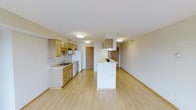 This single-floor space features an open-concept layout that seamlessly connects the kitchen and living area, creating a bright and functional environment. The kitchen is equipped with light wood cabinetry that lines the walls, offering generous storage space alongside under-counter cabinets and drawers. A white refrigerator, range, and built-in microwave provide essential appliances, while the sink is positioned along the counter with a window-adjacent view. A built-in range hood overhead adds a practical touch above the cooking area. A white peninsula extends into the space, serving as a natural divider between the kitchen and the open living area while offering additional counter space or casual seating. Light hardwood flooring runs continuously throughout the entire space, contributing to a clean, cohesive aesthetic. Flush-mount ceiling lights illuminate the interior evenly, maintaining a bright atmosphere. The living area is generously proportioned and open, offering flexible furnishing options. Neutral wall tones and simple trim work keep the overall ambiance clean and understated, while a doorway in the background hints at additional rooms beyond.