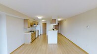 This single-floor space features an open-concept layout that seamlessly connects the kitchen and living area, creating a bright and functional environment. The kitchen is equipped with light wood cabinetry that lines the walls, offering generous storage space alongside under-counter cabinets and drawers. A white refrigerator, range, and built-in microwave provide essential appliances, while the sink is positioned along the counter with a window-adjacent view. A built-in range hood overhead adds a practical touch above the cooking area. A white peninsula extends into the space, serving as a natural divider between the kitchen and the open living area while offering additional counter space or casual seating. Light hardwood flooring runs continuously throughout the entire space, contributing to a clean, cohesive aesthetic. Flush-mount ceiling lights illuminate the interior evenly, maintaining a bright atmosphere. The living area is generously proportioned and open, offering flexible furnishing options. Neutral wall tones and simple trim work keep the overall ambiance clean and understated, while a doorway in the background hints at additional rooms beyond.