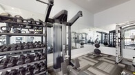 Located at 1779 Central St in Stoughton, Massachusetts, this single-floor fitness center is designed to support a comprehensive workout experience. The space is thoughtfully laid out with distinct zones for both cardio and strength training, all unified by a modern aesthetic of crisp white walls and patterned grey carpet tile flooring.

The cardio area is bathed in natural light, courtesy of large windows that line the walls and look out to the surrounding exterior. Treadmills, a stair climber, a stationary bike, and a rowing machine are among the cardio equipment available. A wall-mounted television and a ceiling fan overhead add comfort to this well-lit zone.

The strength training area features a robust selection of equipment, including a multi-functional cable machine, a Smith machine with a flat bench, and a dedicated free weights section stocked with a full rack of dumbbells and kettlebells in varying sizes. Floor-to-ceiling mirrors span one wall, amplifying the sense of space and allowing users to monitor their form during workouts.

The facility also includes a leg extension machine and additional resistance equipment, rounding out the strength offerings. The high coffered ceiling with recessed lighting in the cardio section adds an architectural polish, while the overall clean, neutral design keeps the focus on functionality and performance.