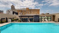 Located in the 70112 zip code of New Orleans, this rooftop pool and terrace offers a well-appointed outdoor retreat set against a striking urban backdrop. The centerpiece is a generously sized swimming pool with vivid turquoise water, framed by clean tiling and bordered by a sleek black metal railing system that provides both safety and unobstructed sightlines to the surrounding cityscape.

Adjacent to the pool, a glass-enclosed pavilion serves as an indoor-outdoor lounge space, constructed with steel framing and floor-to-ceiling glazing that allows natural light to flood the interior. The pavilion features recessed lighting and connects seamlessly to the open terrace through stainless steel-framed glass doors, facilitating easy transitions between the interior and exterior zones.

The open terrace areas are finished with large-format stone pavers and composite wood decking, providing distinct zones for relaxation. Plush wicker sectional sofas with neutral cushions are positioned throughout, offering comfortable seating with views of the New Orleans skyline. Raised planters filled with lush greenery add a natural element to the rooftop environment. Staircase access, perimeter fencing, and thoughtful landscaping complete this elevated urban outdoor space.